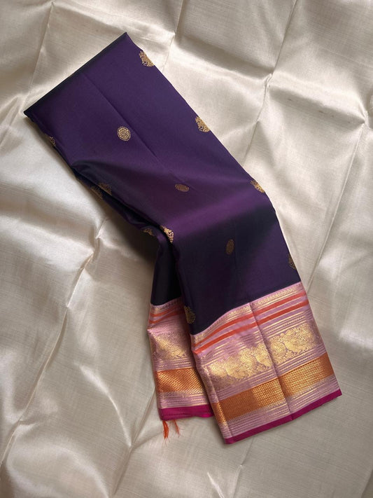 Shyamala—named after the deep, dark shade it embodies 💜 A pure Kanchipuram silk masterpiece with a rich purple body sprinkled with golden zari buttas. The contrast pink border glows with floral and paisley zari motifs, accented in coral and rose-gold, wh