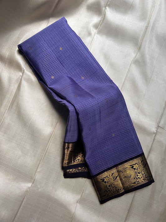 Classic heritage finesse in this lilac blue pure Kanchipuram silk saree Zari kattam , intricately handwoven with fine golden checks across the body. Scattered floral buttas add delicate charm,rich purple peacock border