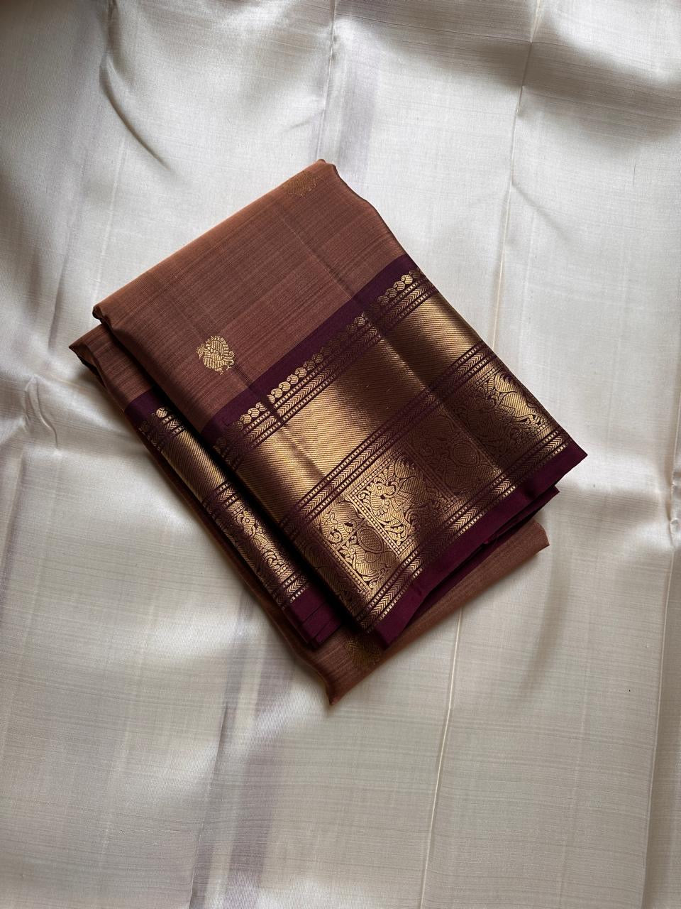 Pari is a graceful Kanchipuram pure silk saree in a serene copper-rose gold brown hue. Scattered peacock zari buttas .  Framed by an antique-gold korvai border temple-inspired yali, annam, and floral medallions, finished with a deep wine-maroon Pallu