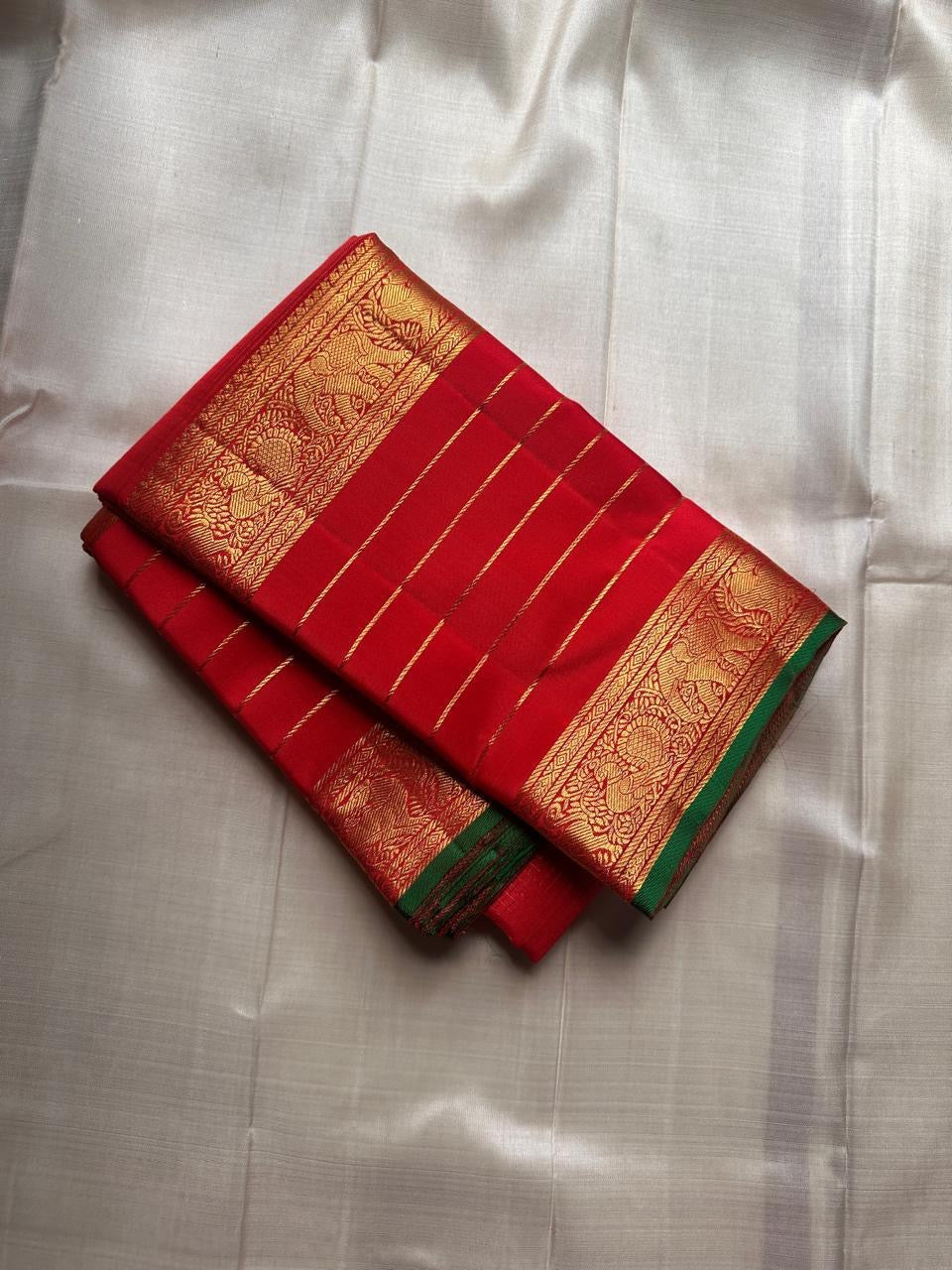 Folded red Kanchipuram silk saree with gold horizontal zari lines, rich elephant border and green selvedge on an ivory background.