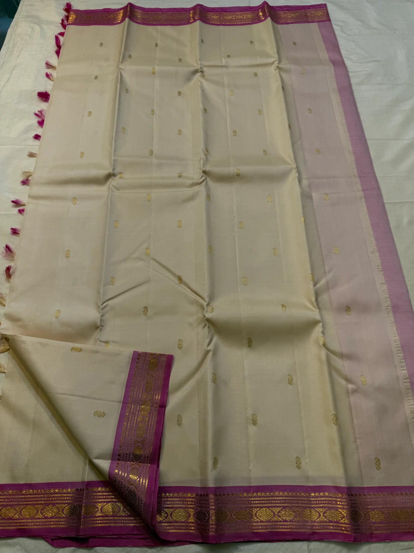 Full-length cream silk saree laid flat with scattered gold buttas, magenta gold-zari border, and a light lavender side panel.