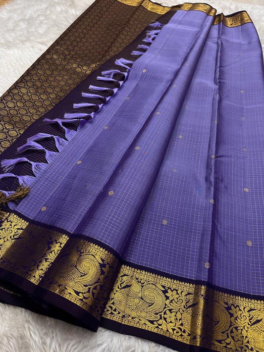 Classic heritage finesse in this lilac blue pure Kanchipuram silk saree Zari kattam , intricately handwoven with fine golden checks across the body. Scattered floral buttas add delicate charm,rich purple peacock border