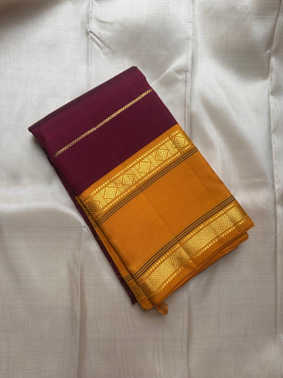 Folded deep maroon pure Kanchipuram silk saree with mustard gold zari border, shown on a light fabric background.