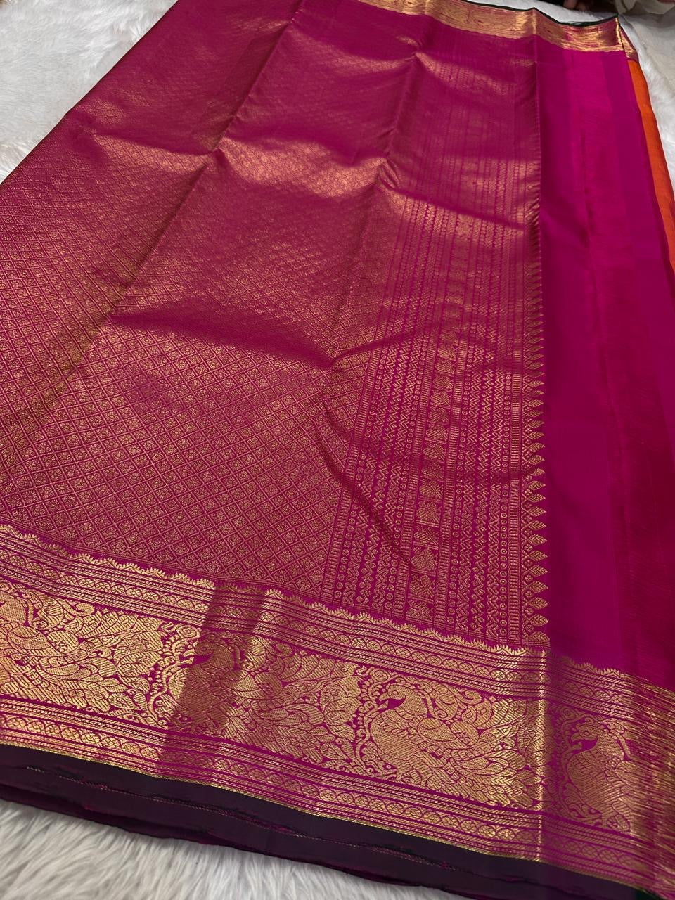 Sharvani – Orange & Magenta Pure Silk Kanchipuram Saree
Featuring zari-woven stripes across the body for subtle structure.
Bold magenta border with rice reed and peacock motifs, finished with a green selvedge.