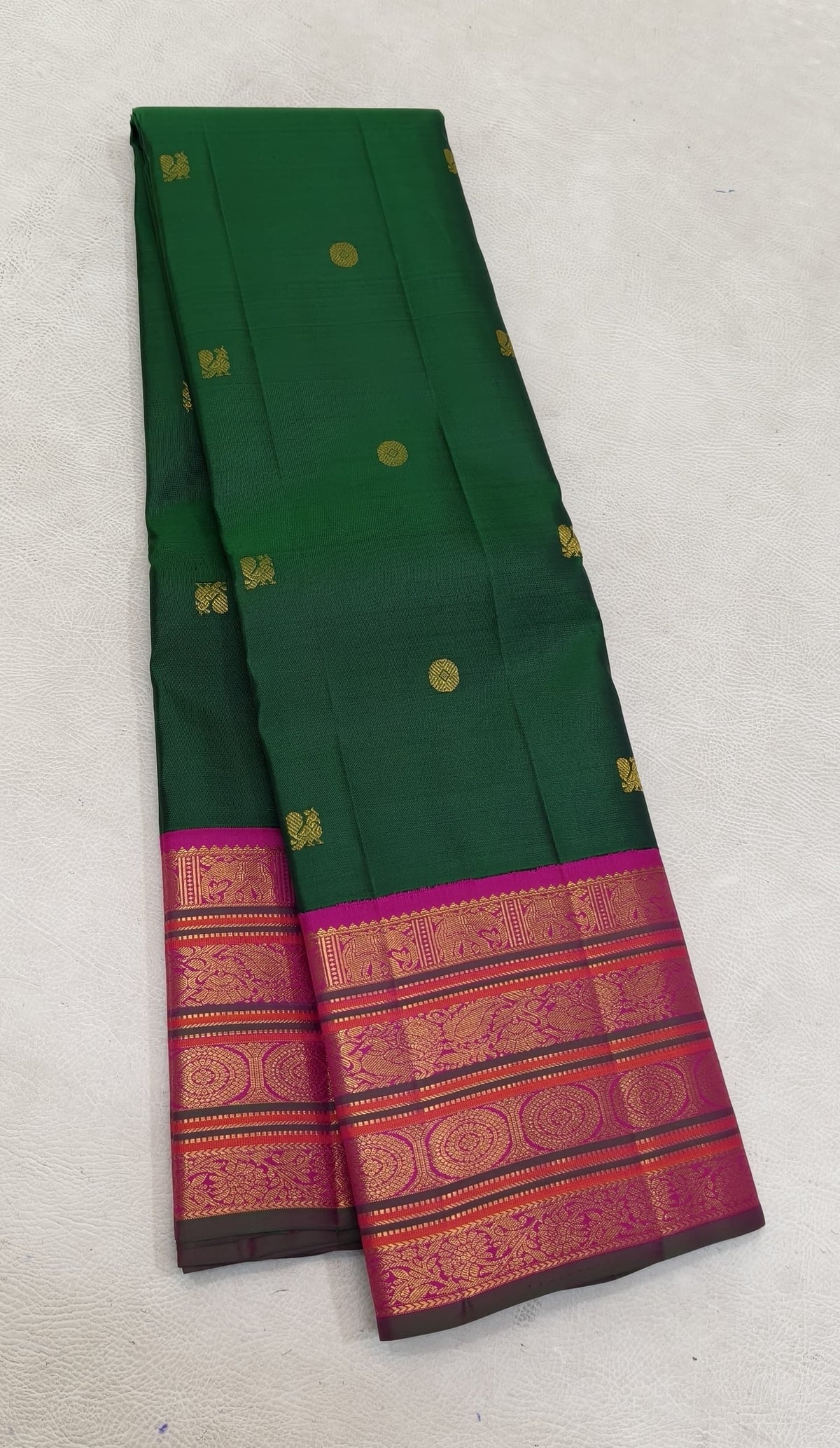 Folded deep emerald-green Kanchipuram silk saree with scattered gold zari buttas and a vivid magenta Varisaipet border.