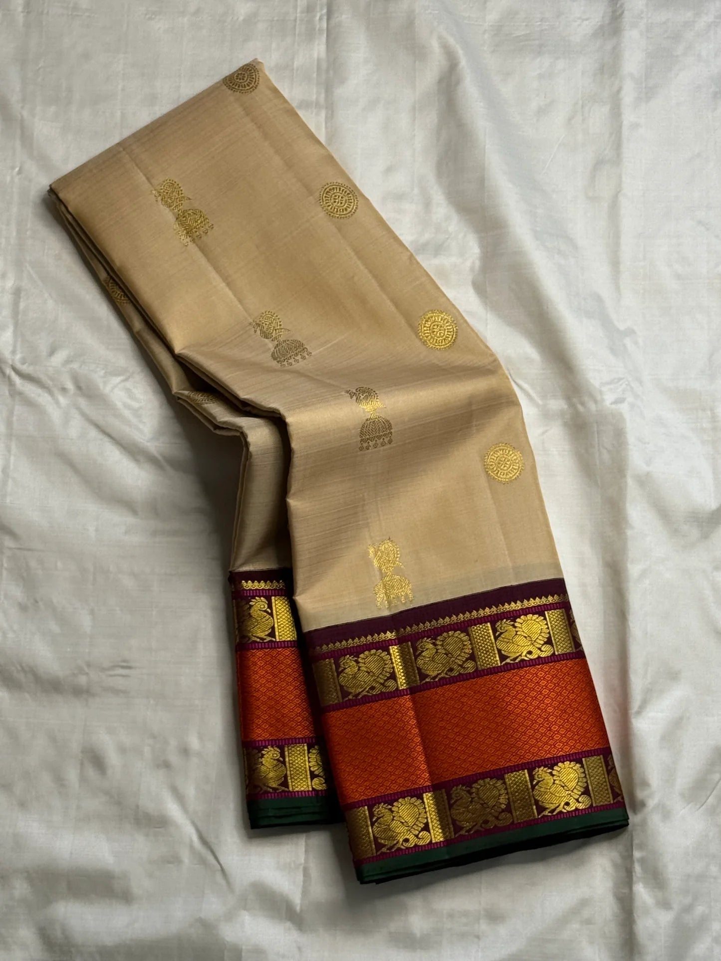 Biscuit beige pure Kanchipuram silk saree draped to show gold buttas and wide rust orange zari woven border with maroon detailing and green selvedge