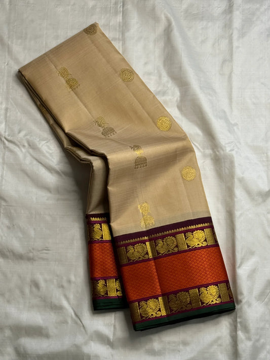 Biscuit beige pure Kanchipuram silk saree draped to show gold buttas and wide rust orange zari woven border with maroon detailing and green selvedge