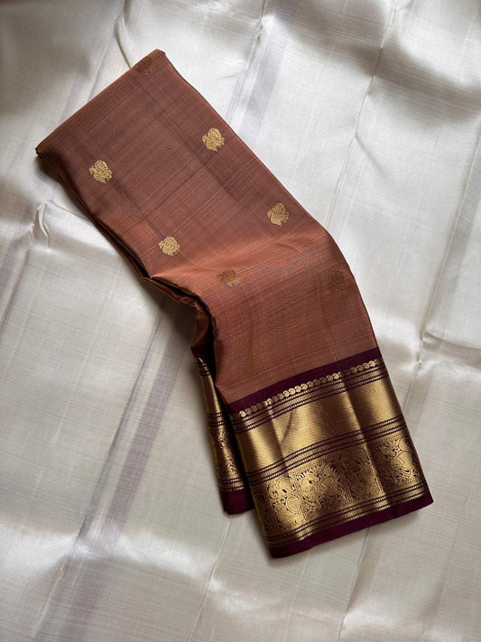Pari is a graceful Kanchipuram pure silk saree in a serene copper-rose gold brown hue. Scattered peacock zari buttas .  Framed by an antique-gold korvai border temple-inspired yali, annam, and floral medallions, finished with a deep wine-maroon Pallu