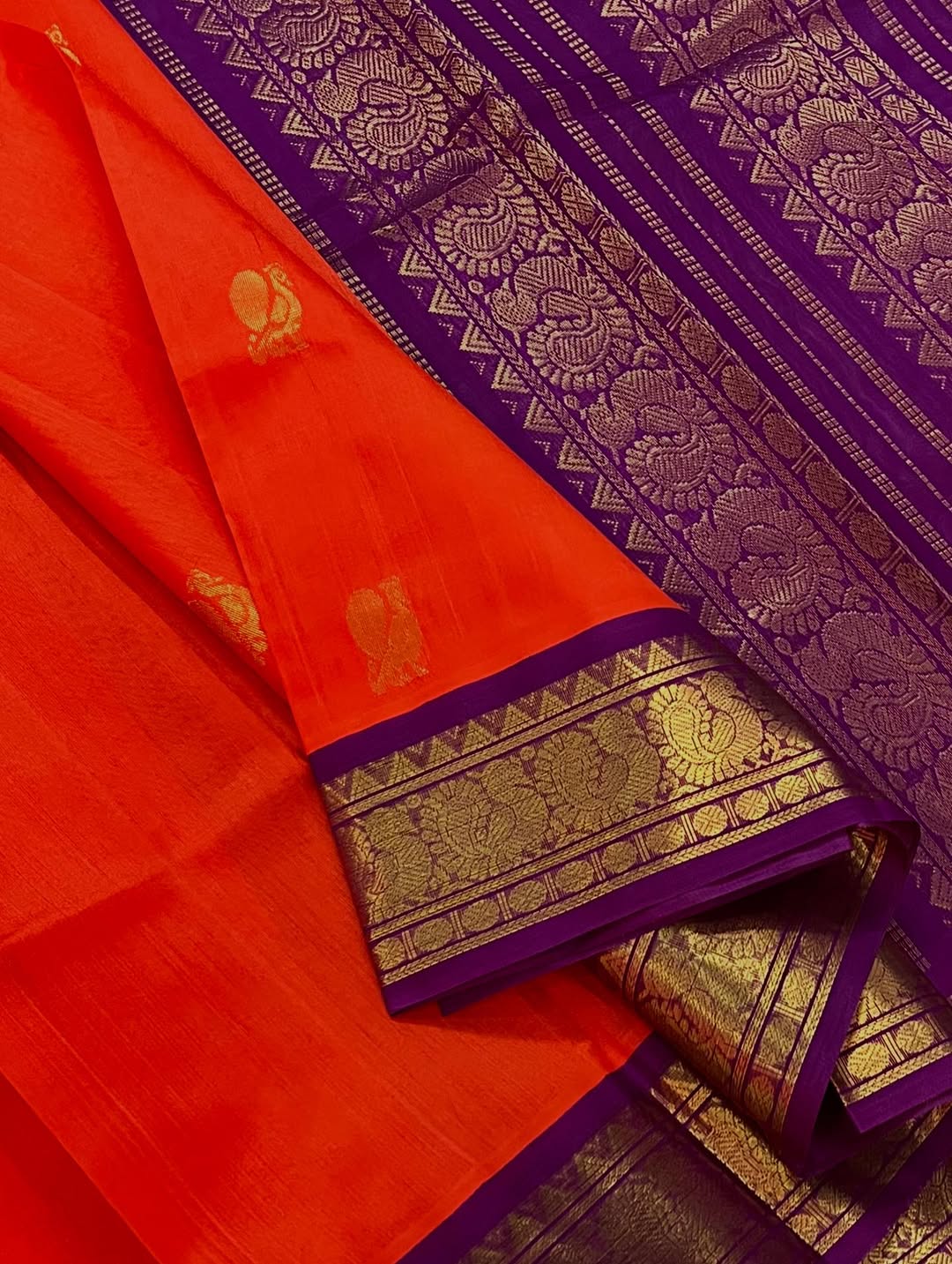 An exquisite blend of tradition and vibrancy, this silk cotton saree features a radiant orange body adorned with elegant gold zari buttas, The rich contrast border and pallu in royal purple and intricately woven with traditional gold zari motif