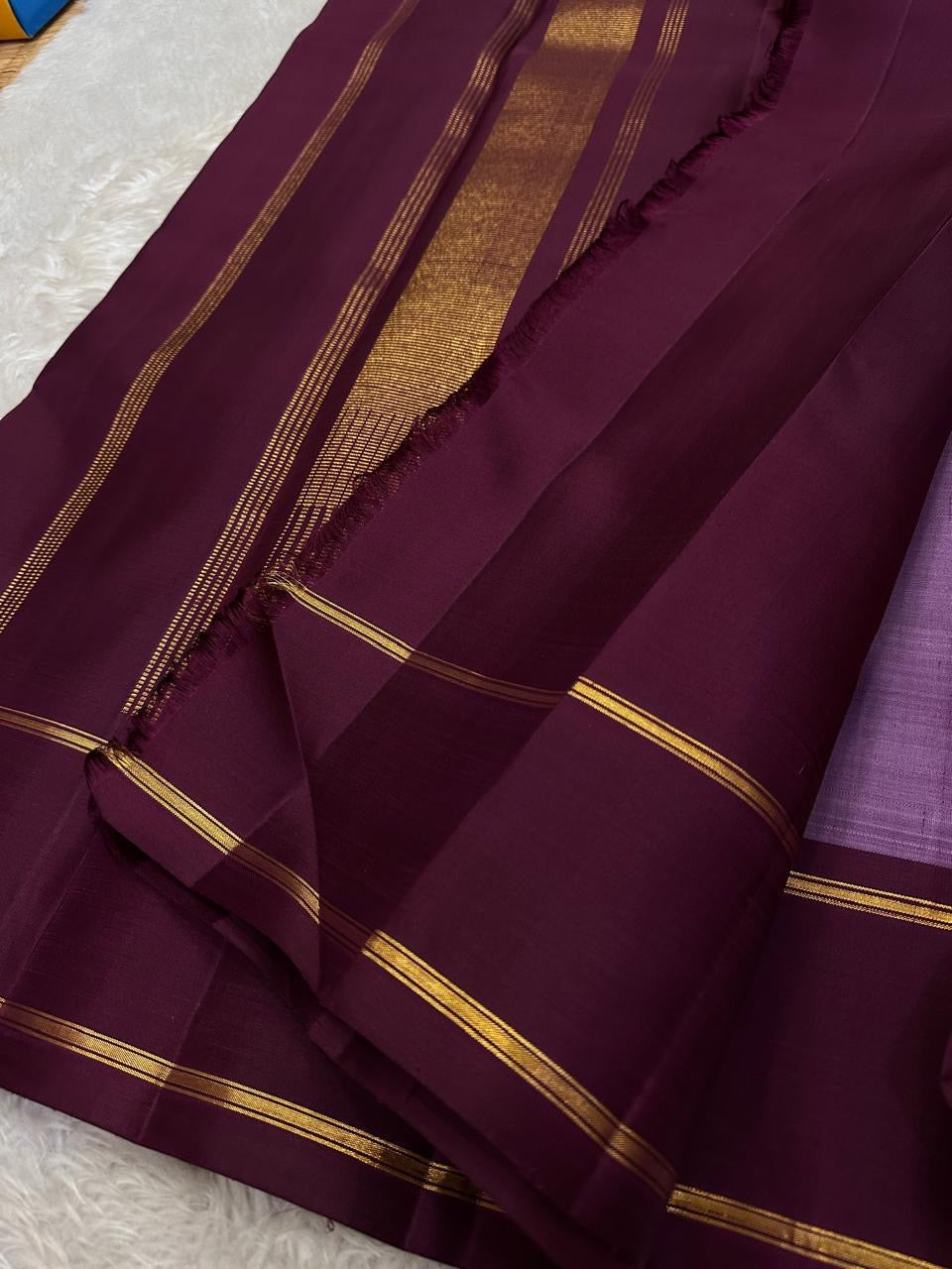 Ilam  Vadamalli Chakram — Kanchipuram Pure Silk

Pastel lilac–mauve body with gold & silver chakram buttas

Deep plum contrast border with twin kaddi zari lines

Plum pallu with elegant vertical zari stripes; lilac tassels

Silk Mark Certified •
