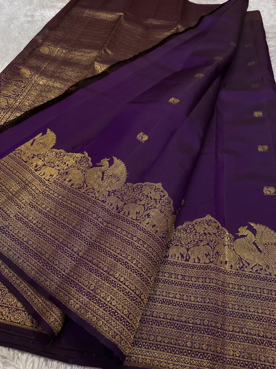 Swarna
Majestic Purple Pure Silk Kanchipuram Saree with Broad Varisaipet Zari Border,Intricately Woven Peacock and Elephant Motifs alongside border . Peacock motifs all over.