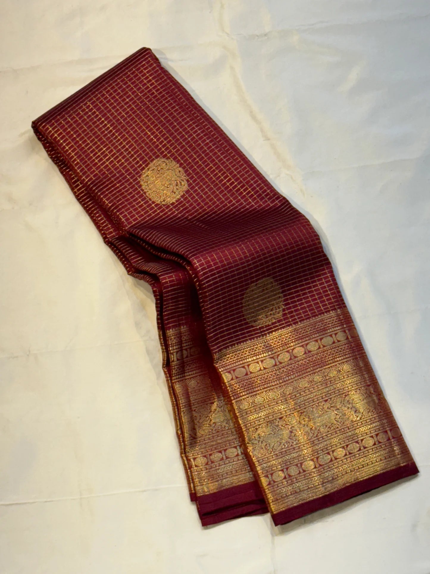 Pavila – Rich brown pure silk Kanchipuram saree in shimmering zari kattam checks. Layered varisaipet borders feature peacocks, yazhi, and rudraksham motifs in ornate zari. A bold broad yazhi adai motif lends ceremonial elegance. Grand  ornate Pallu