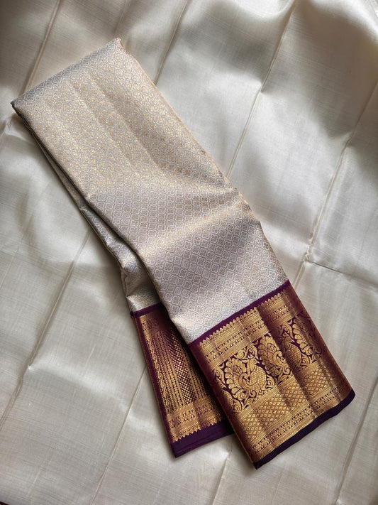 Ivory silk meets tradition in this elegant Kanchipuram saree. With a deep purple border and Mayuri motifs, it’s perfect for moments that call for quiet richness.