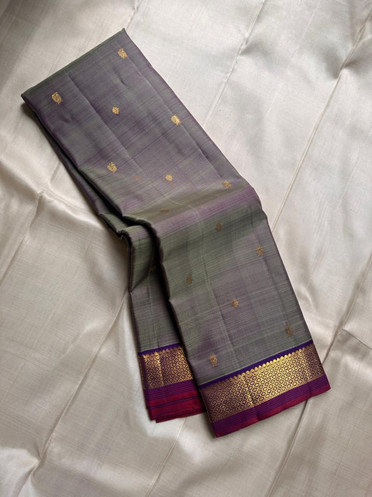 Varnam – Dove Tail Gray & Purple, a Kanchipuram silk saree where elegance meets bold expression. The soft dove tail gray base is adorned with vibrant magenta checks,Framed by a rich purple zari border and finished with an intricate contrast pallu