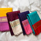 Assorted pure Kanchipuram silk sarees in yellow, magenta, maroon, cream, purple, pink, and Rama green, arranged side by side.
