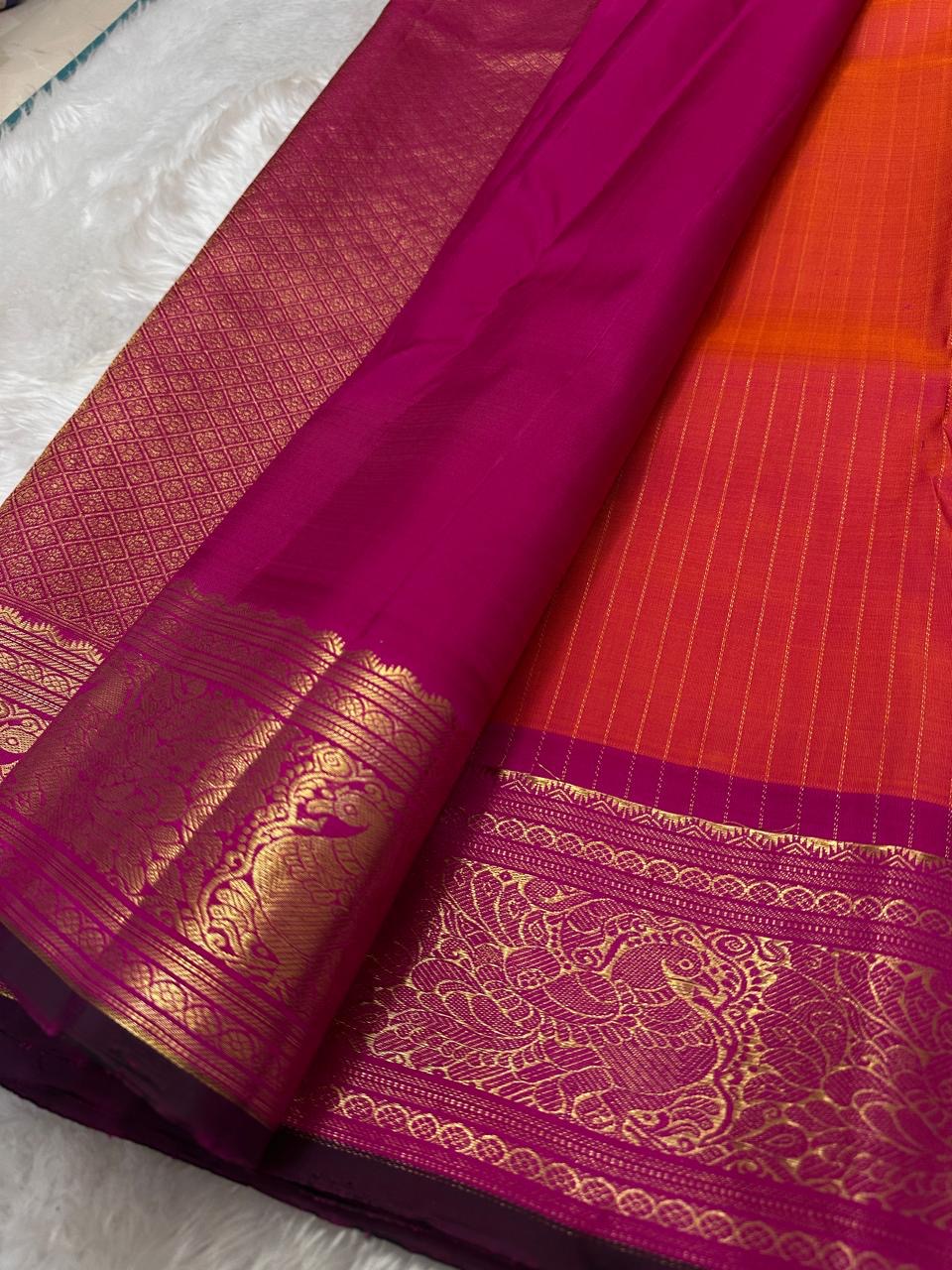 Sharvani – Orange & Magenta Pure Silk Kanchipuram Saree
Featuring zari-woven stripes across the body for subtle structure.
Bold magenta border with rice reed and peacock motifs, finished with a green selvedge.