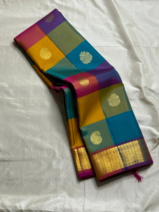 Rangoli Kattam – Pure Silk Kanchipuram Saree
Vibrant multicolor kattam checks with golden zari buttas across the body
Rich rani pink pallu with intricate zari weaving for a grand finish
Contrast purple borders edged with gleaming zari motifs