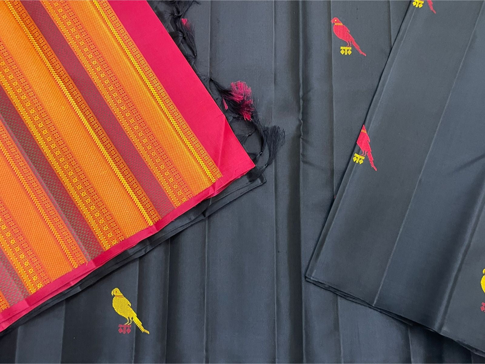 Close-up of orange striped pallu with black tassels beside black Pure Kanchipuram Silk Saree fabric with a yellow bird motif, shipped from New Jersey USA.