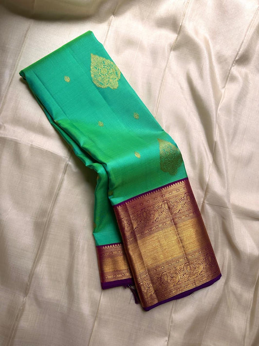 This pure silk Kanchipuram saree features a radiant shot color blend of green, gold, and blue, adorned with delicate zari buttas. A regal royal purple rettapet border and an elaborate peacock motif pallu complete this timeless drape — perfect for festive