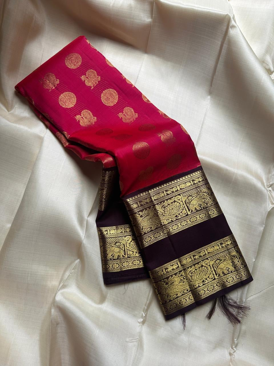 A regal  shot tomato red pink Kanchipuram silk saree features elegant chakram and mayil gold zari buttas woven across the body. Brown contrast rettapet border and Pallu vanasringaram details