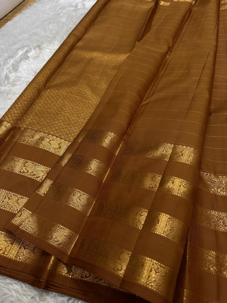 vasantham  Brown– Handwoven Kanchipuram Silk Saree A poem in Brown silk. The body glows in a serene earthy Brown, softly accented by delicate gold vertical lines and dainty gold zari buttas that glint like little blessings.