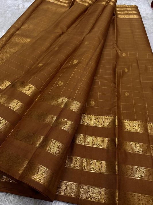 vasantham  Brown– Handwoven Kanchipuram Silk Saree A poem in Brown silk. The body glows in a serene earthy Brown, softly accented by delicate gold vertical lines and dainty gold zari buttas that glint like little blessings.