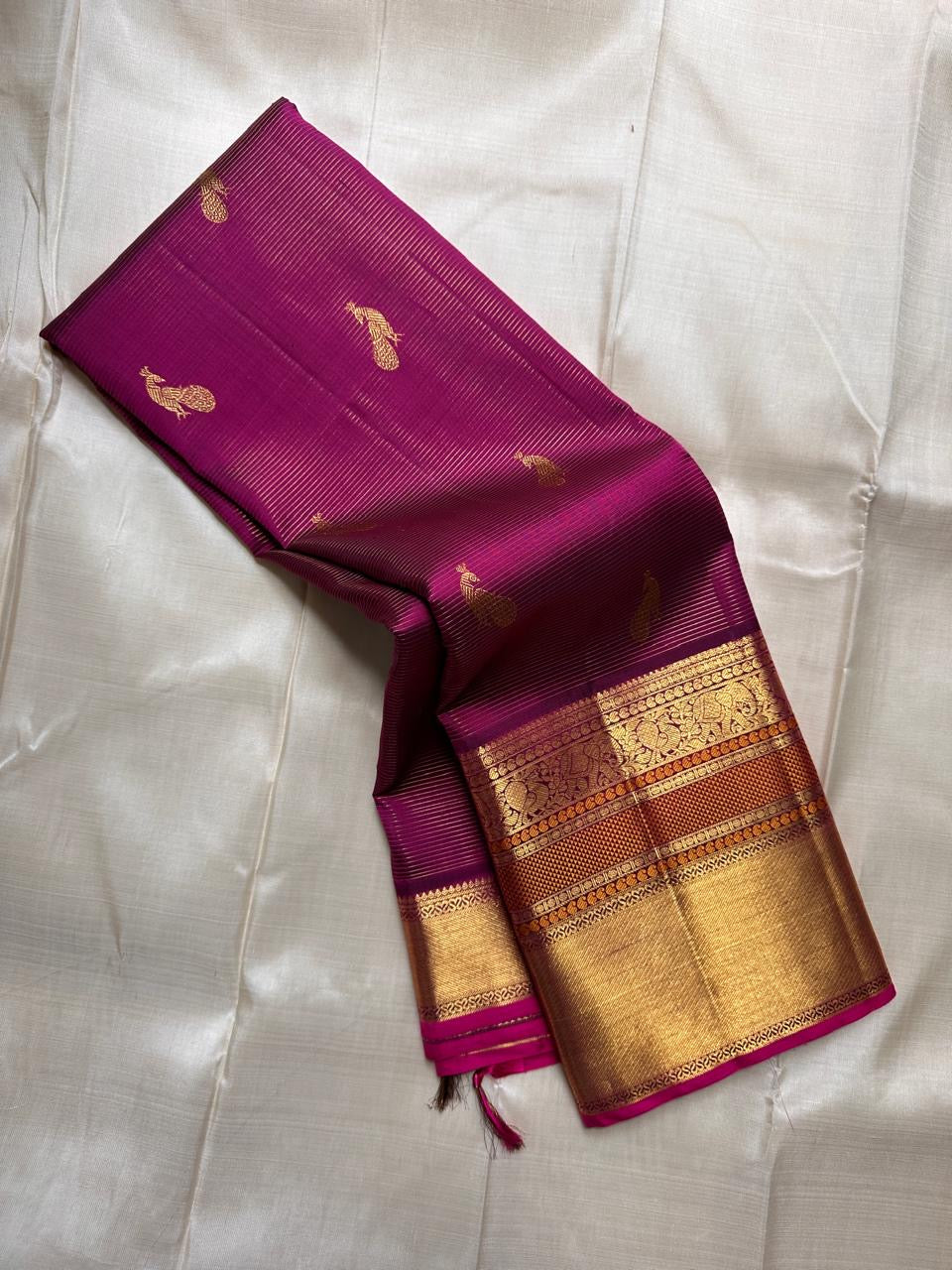Folded magenta Kanchipuram silk saree with gold peacock buttas and rich zari border on an ivory backdrop
