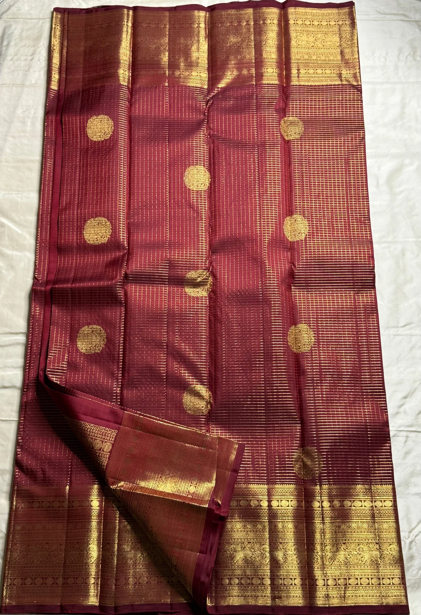 Pavila – Rich brown pure silk Kanchipuram saree in shimmering zari kattam checks. Layered varisaipet borders feature peacocks, yazhi, and rudraksham motifs in ornate zari. A bold broad yazhi adai motif lends ceremonial elegance. Grand  ornate Pallu
