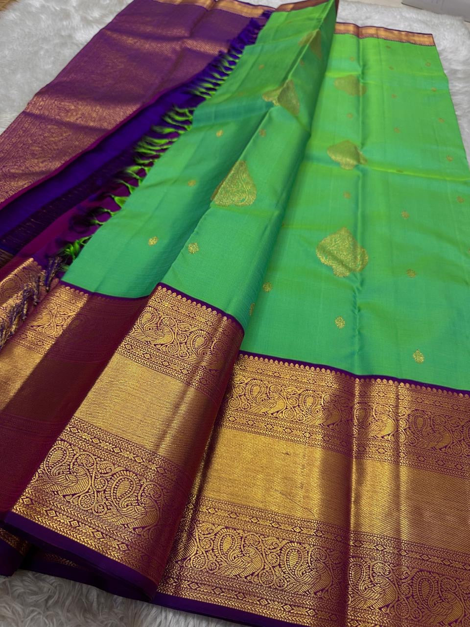 This pure silk Kanchipuram saree features a radiant shot color blend of green, gold, and blue, adorned with delicate zari buttas. A regal royal purple rettapet border and an elaborate peacock motif pallu complete this timeless drape — perfect for festive
