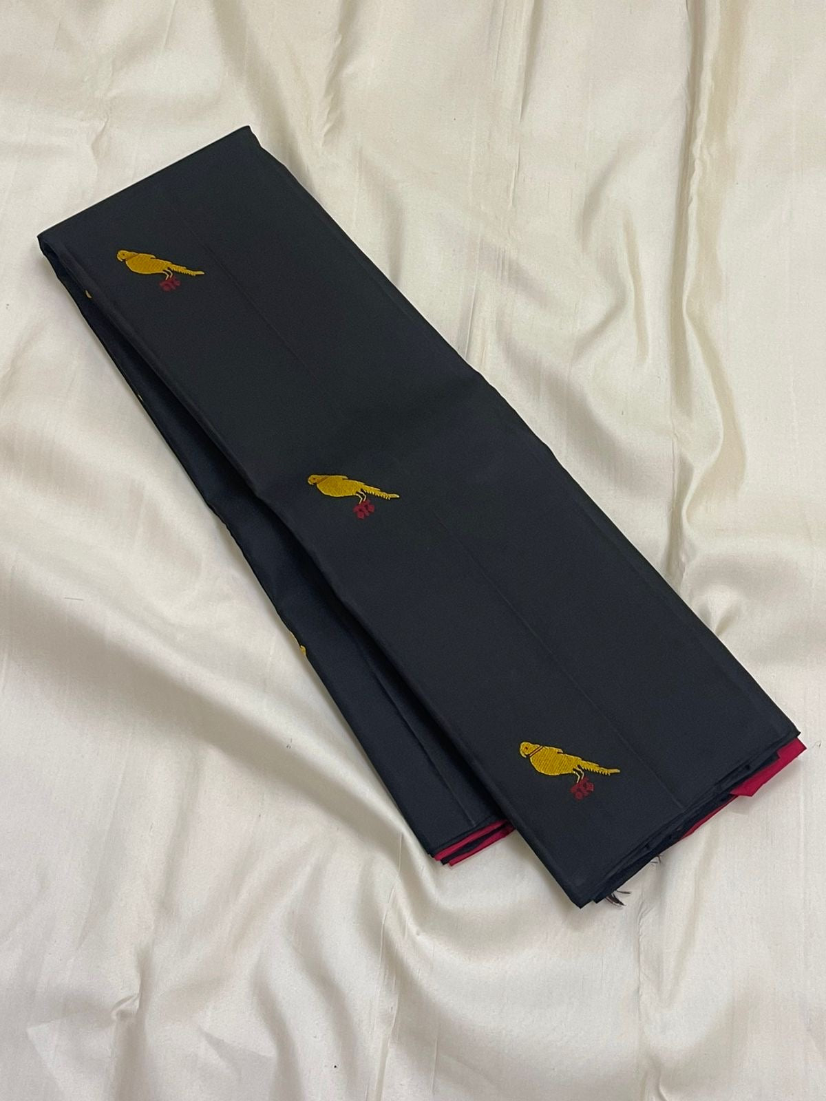 Long folded black Pure Kanchipuram Silk Saree on white fabric, showing scattered yellow bird motifs and a pink edge