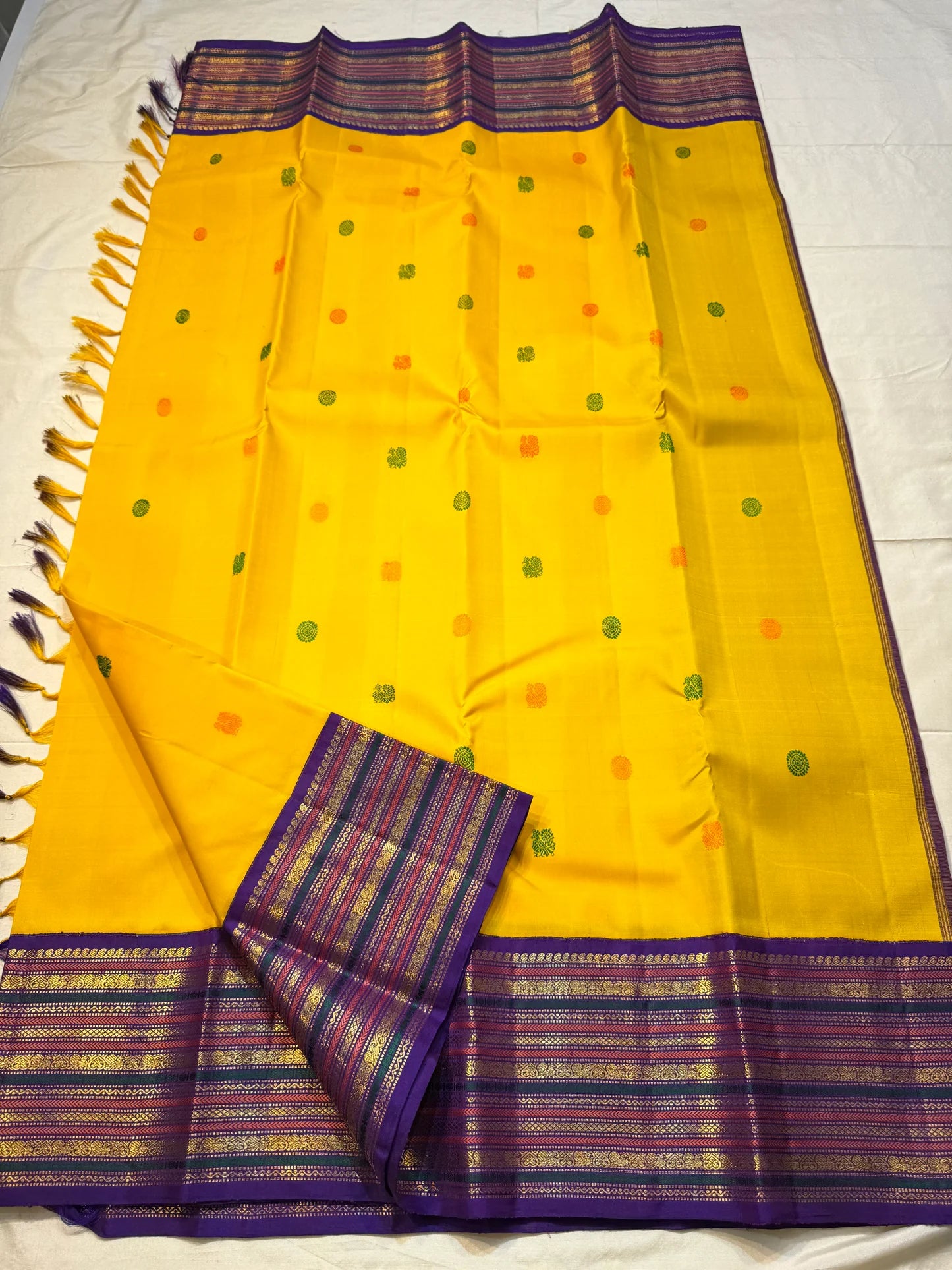 Swarna Kanakam Mayil Chakram — Pure Kanchipuram Silk Saree
Radiant turmeric-gold body adorned with silk-thread mayil and chakram buttas, framed by a regal vadamalli zari-woven korvai border and rich purple pallu.
Silk Mark Certified • Blouse Stitching Ava