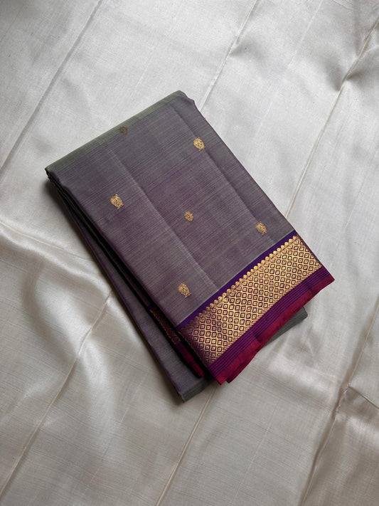 Varnam – Dove Tail Gray & Purple, a Kanchipuram silk saree where elegance meets bold expression. The soft dove tail gray base is adorned with vibrant magenta checks,Framed by a rich purple zari border and finished with an intricate contrast pallu