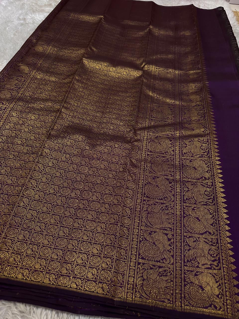 Swarna
Majestic Purple Pure Silk Kanchipuram Saree with Broad Varisaipet Zari Border,Intricately Woven Peacock and Elephant Motifs alongside border . Peacock motifs all over.