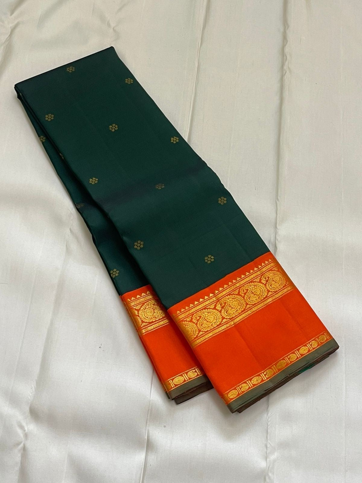 Pachai Kesar Mayil — Kanchipuram Pure Silk

 Bottle-green body with dainty gold floral buttas

Contrast marigold-orange korvai borders with temple edging

Rich gold-zari paisley scrolls; striped orange pallu with tassels

Silk Mark Certified | Free Shipp