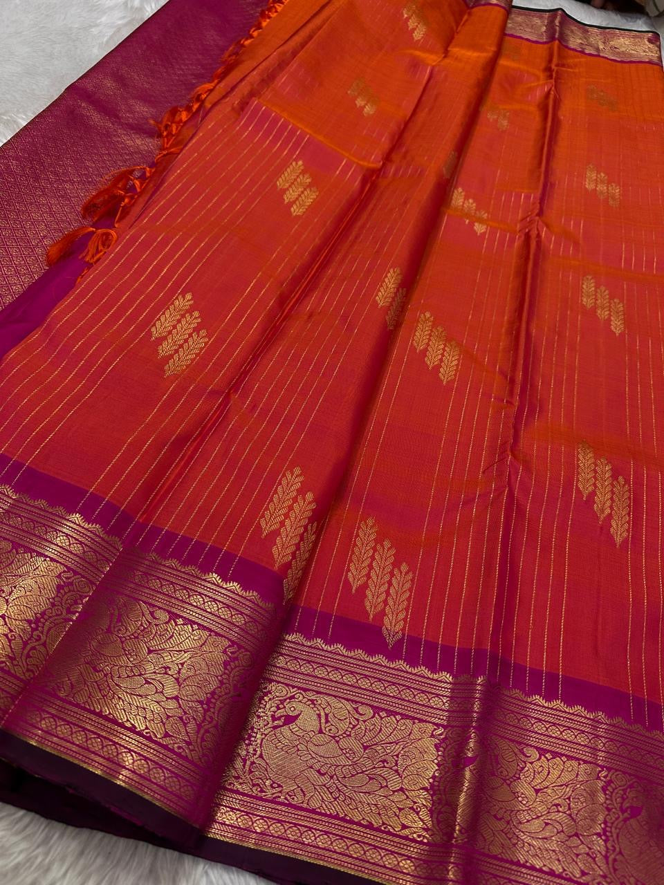 Sharvani – Orange & Magenta Pure Silk Kanchipuram Saree
Featuring zari-woven stripes across the body for subtle structure.
Bold magenta border with rice reed and peacock motifs, finished with a green selvedge.