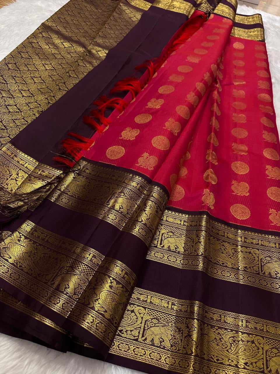 A regal  shot tomato red pink Kanchipuram silk saree features elegant chakram and mayil gold zari buttas woven across the body. Brown contrast rettapet border and Pallu vanasringaram details