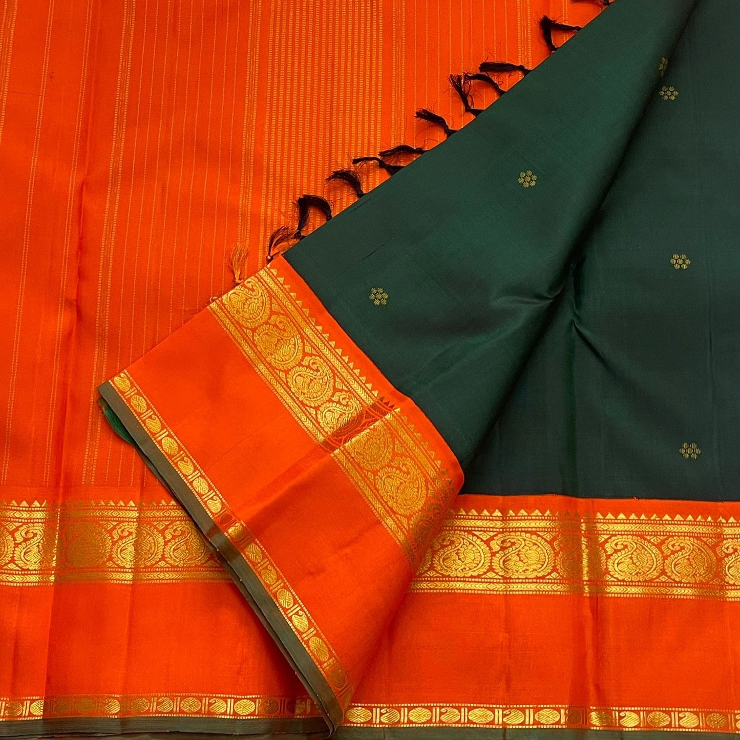 Pachai Kesar Mayil — Kanchipuram Pure Silk

 Bottle-green body with dainty gold floral buttas

Contrast marigold-orange korvai borders with temple edging

Rich gold-zari paisley scrolls; striped orange pallu with tassels

Silk Mark Certified | Free Shipp