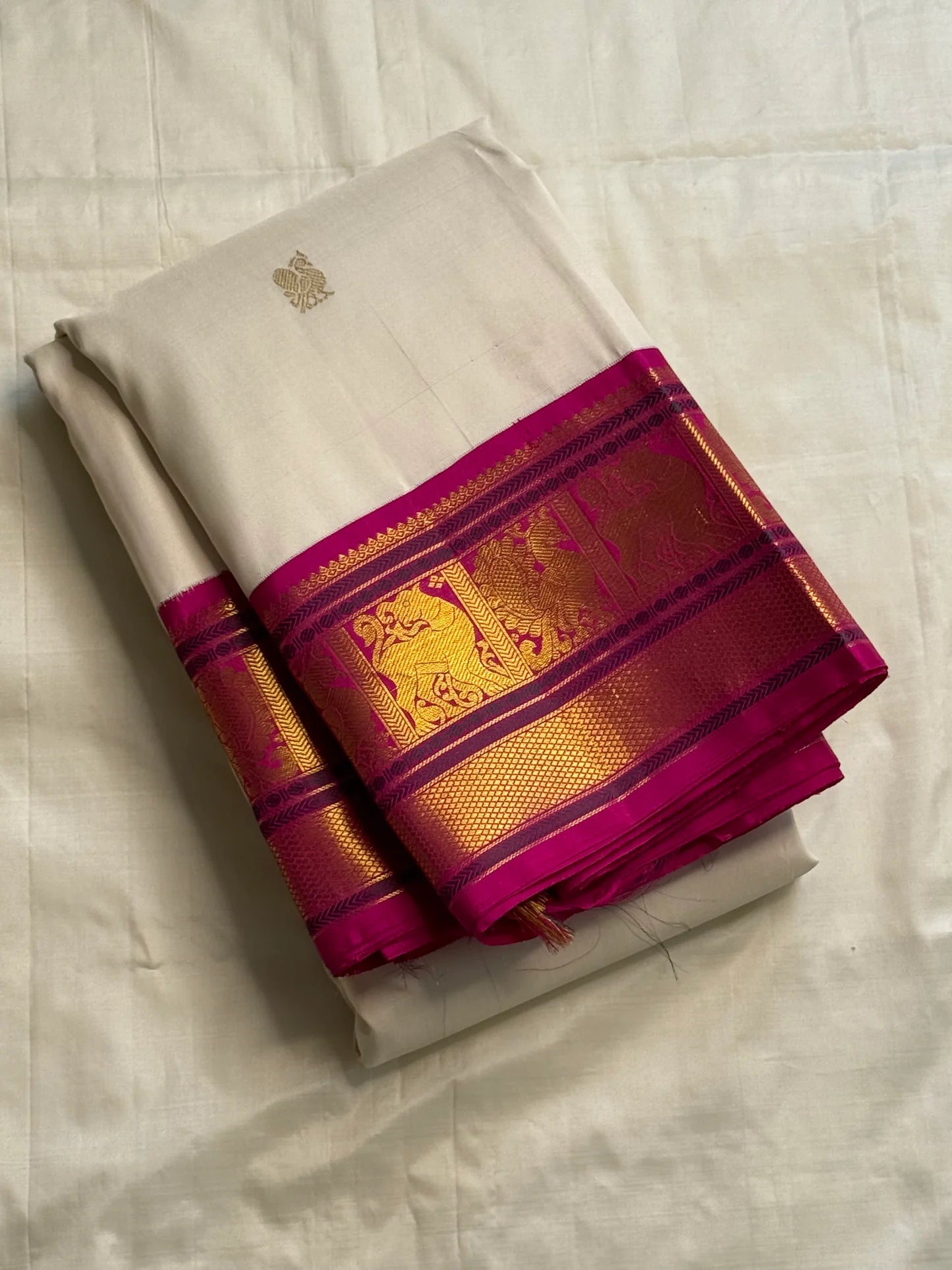 Folded ivory silk saree with fuchsia border and gold zari woven panels, photographed on a white fabric background.