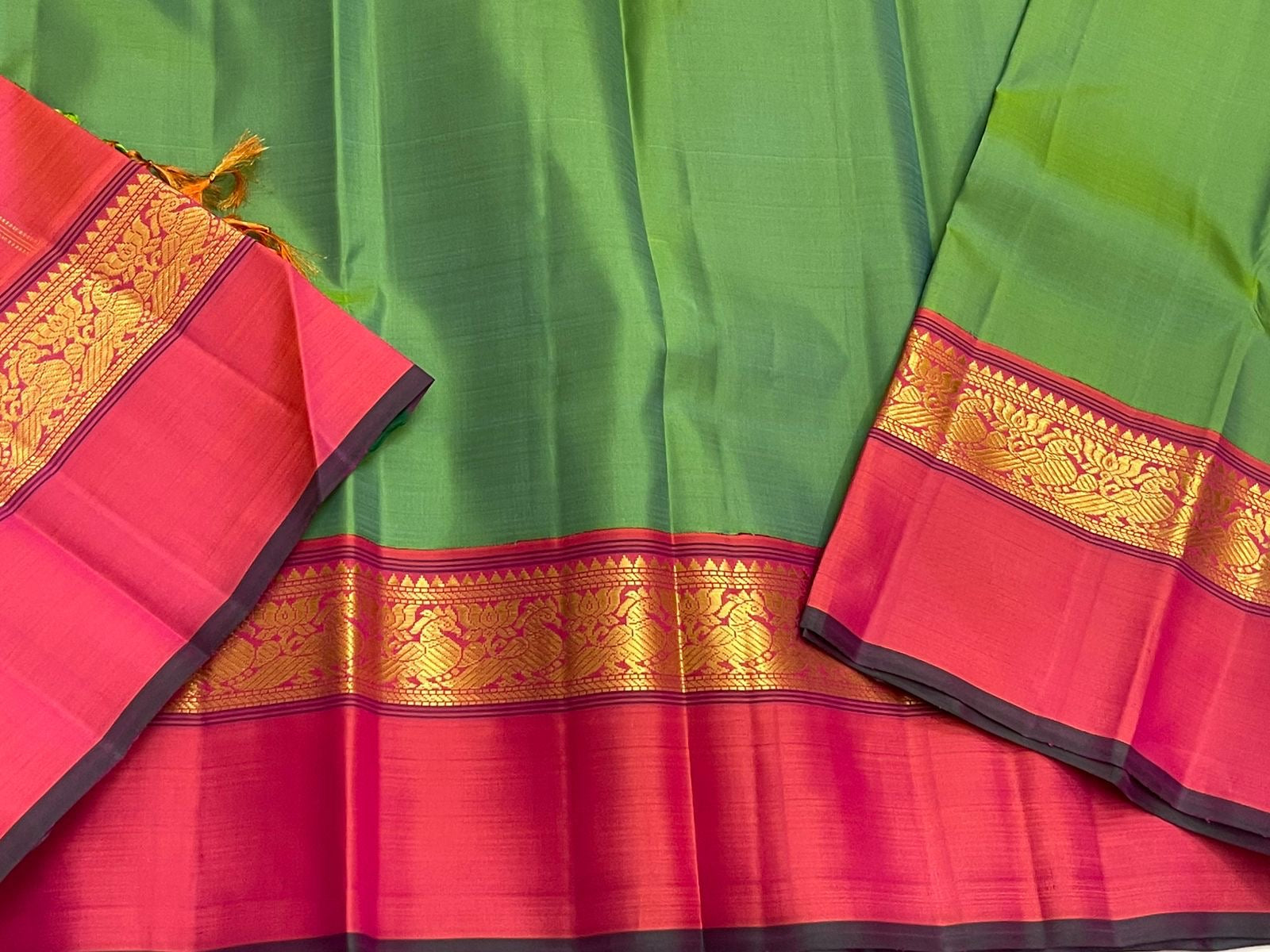 Close-up of green saree with wide pink border and gold zari woven peacock band, with dark piping on the edge.