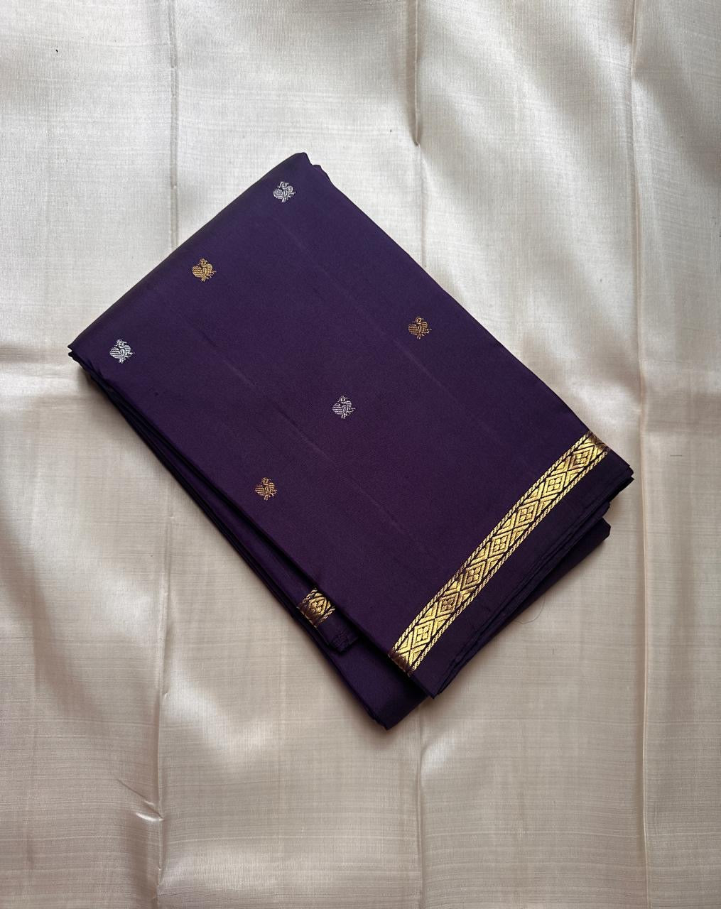 Folded deep purple Kanchipuram silk saree with small gold and silver buttas and slim diamond zari border on an ivory background.