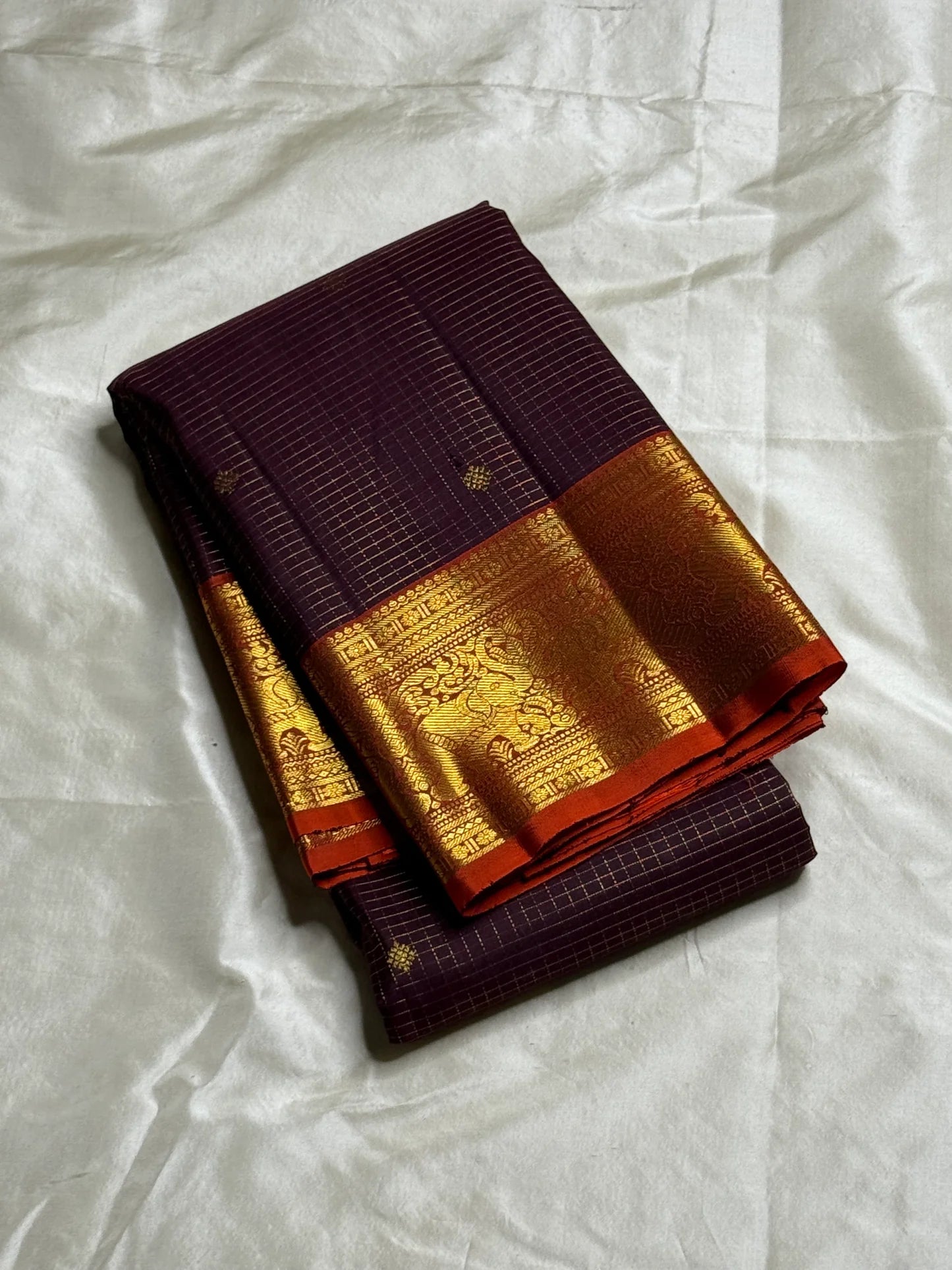 Deep Plum Orange
A regal dance of plum and burnt orange
Micro-checks kissed with golden buttas
Elephant borders marching in heritage grandeur
Brocade pallu glowing in timeless zari brilliance

Silk Mark Certified, delivery in 2 weeks
