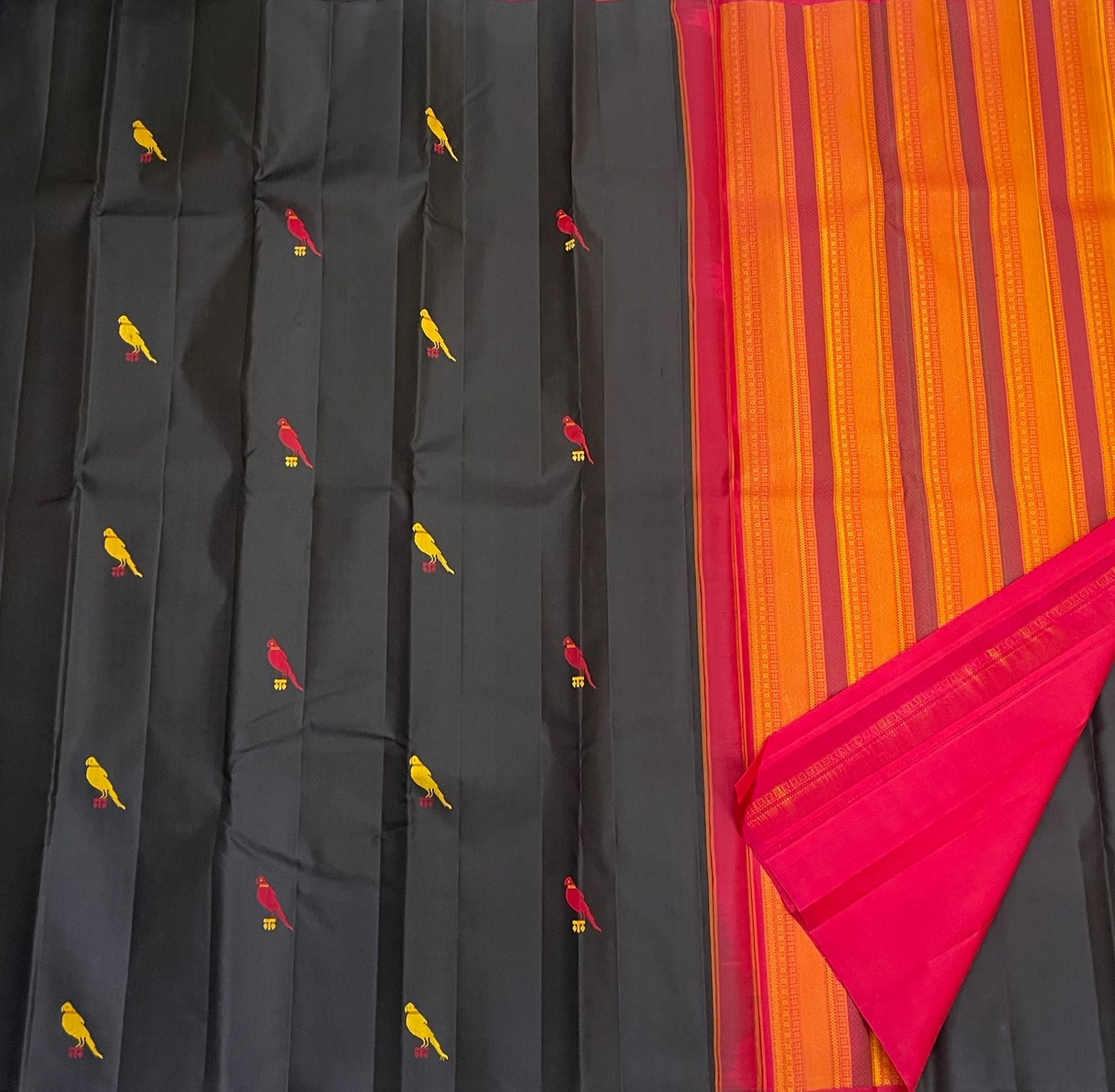 Flat lay of black Pure Kanchipuram Silk Saree with yellow and red bird motifs next to an orange striped pallu and pink border, available in the USA.