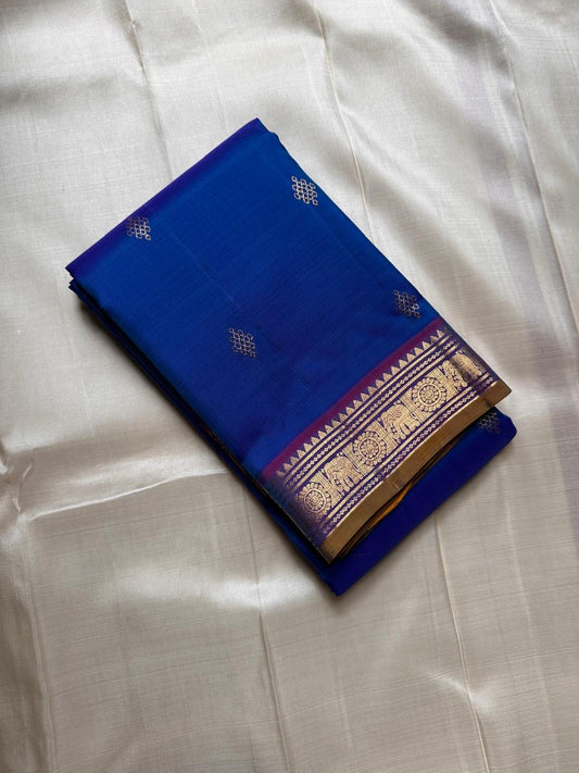 Sapphire Orchid celebrates the classic blue–purple pairing of Kanchi. The body is a rich sapphire/cobalt blue with tiny floral buttas in gold zari. A striking amethyst-purple korvai border and grid pavun Zari pallu Silk Mark Certified, delivery in 2 weeks