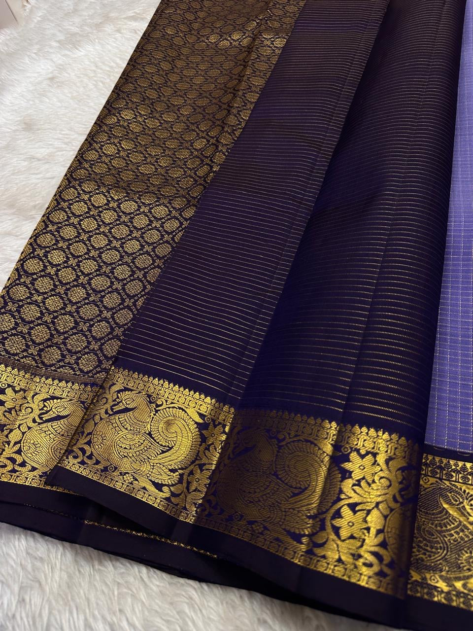 Classic heritage finesse in this lilac blue pure Kanchipuram silk saree Zari kattam , intricately handwoven with fine golden checks across the body. Scattered floral buttas add delicate charm,rich purple peacock border