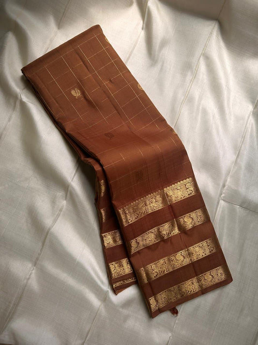 vasantham  Brown– Handwoven Kanchipuram Silk Saree A poem in Brown silk. The body glows in a serene earthy Brown, softly accented by delicate gold vertical lines and dainty gold zari buttas that glint like little blessings.