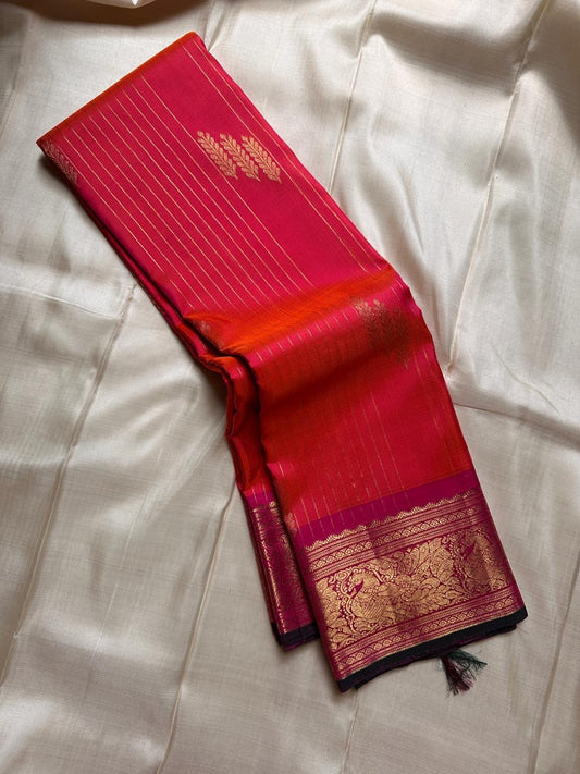 Sharvani – Orange & Magenta Pure Silk Kanchipuram Saree
Featuring zari-woven stripes across the body for subtle structure.
Bold magenta border with rice reed and peacock motifs, finished with a green selvedge.