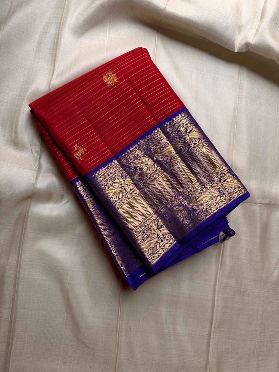 Raaga – Red and Blue Pure Kanchipuram Silk Saree with Gold Zari Border
Features a striped red body with small gold buttas and a royal blue pallu.
Finished with intricately woven peacock motifs in pure zari across the border.