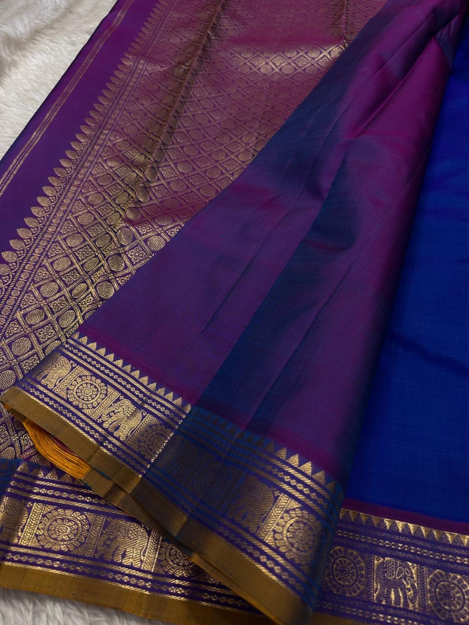 Sapphire Orchid celebrates the classic blue–purple pairing of Kanchi. The body is a rich sapphire/cobalt blue with tiny floral buttas in gold zari. A striking amethyst-purple korvai border and grid pavun Zari pallu Silk Mark Certified, delivery in 2 weeks