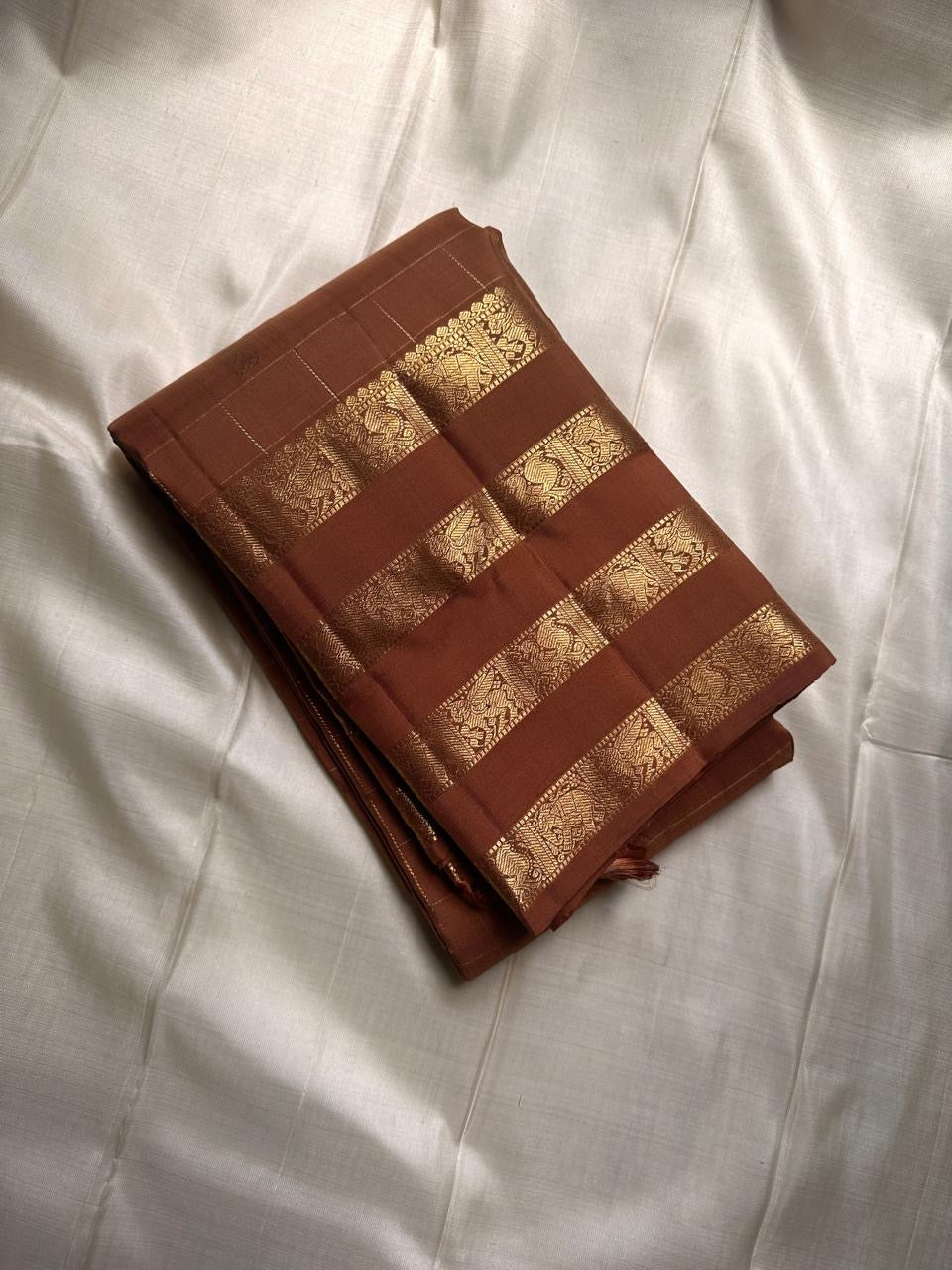vasantham  Brown– Handwoven Kanchipuram Silk Saree A poem in Brown silk. The body glows in a serene earthy Brown, softly accented by delicate gold vertical lines and dainty gold zari buttas that glint like little blessings.