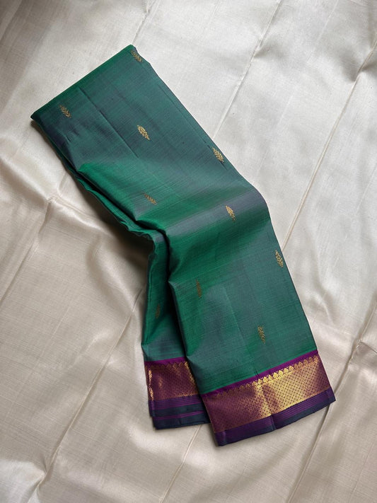 Varnam is a pure Kanchipuram silk saree in rich emerald green with subtle dual-tone shading. Woven gold zari buttas dot the body, while a petite purple border woven with intricate zari adds refined contrast. Arai maadam Pallu, jewel toned classic !